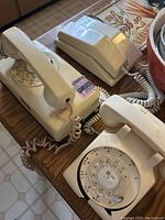 Photo showing three vintage rotary telephones in beige: two hanging-style with metal hooks and one table-style phone, all with rotary dials and coiled cords.