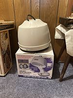 Large dome-shaped vintage hair dryer on box with another boxed vintage 1200 watt salon hair dryer, placed on carpeted floor against wood paneling