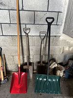 Photo showing two metal shovels with square tips and D-handles, a red scoop shovel with wooden handle, and a green plastic snow shovel with metal handle leaning against a concrete wall.