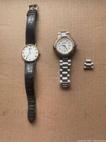 Two men's wristwatches side by side on a surface. Left watch is a Seiko with a black leather band; right watch is a Guess with a silver-tone linked metal band and an additional removable link beside it.