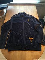 Full view of the front of the midnight blue velour jacket with yellow piping and embroidered logos, laid flat on wooden table.