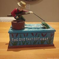 Side view showing the fisherman figure aiming a fishing pole at a fish jumping out of the water beside a bucket on a blue base with painted reeds and text.