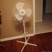 Full view of the white pedestal fan standing on a wood floor against a beige wall, showing the adjustable height stand and protective cage around the blades.
