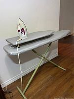 Proctor & Silex electric iron placed on large ironing board with collapsible legs and grey cover, positioned against a light wall and wood floor.