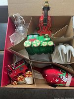 Top view of box with red violin ornament, green train ornament, red locomotive, white deer pair, and red elongated ornament with snowman graphic.