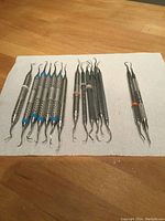 Full view of 15 stainless steel dental instruments arranged on white cloth on wooden table, showing different tip designs and colored bands on handles