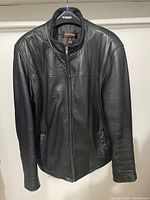 Front view of the black leather jacket hanging on a hanger, zip-up front, two exterior pockets visible.
