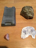Photo showing the 200 gram zinc bar with clear embossed text, metallic ore chunk with yellow patches, quartz slice, and polished bloodstone on wooden surface