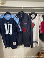 Three NFL team apparel items hanging together on a rack: Tennessee Titans jersey, Buffalo Bills jacket, Atlanta Falcons jersey.