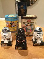 Photo of two novelty tin cans (Looney Tunes and Tootsie Roll) behind 3 Star Wars noise makers (2 R2-D2 and 1 Darth Vader) characters on a table with E.T. VHS cassette in background.