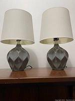 Pair of lamps with grey geometric ceramic bases and white shades on wooden table against white wall.
