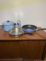 Three cookware items: tall blue enamel pot with lid, stainless steel frying pan, and blue Teflon frying pan shown with water bottle for scale