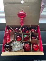 Full view of box containing various red glass Christmas ornaments including large hand-blown shapes and traditional round baubles