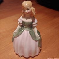 Front angled view of the Royal Doulton Penny figurine on a wooden surface, showing the pink and green dress and blonde hair details.