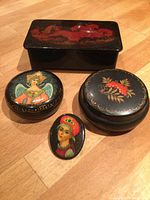 Four items on wooden surface: three Russian-style hand-painted lacquer boxes and one pin. Black rectangular box with red horse on lid, two round boxes with traditional artwork, and a painted pin of woman's portrait.