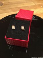 Pair of small rectangular tri-coloured gold pierced earrings displayed on a black surface inside a red box with black interior.