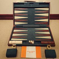 Open backgammon game showing board with green suede playing surface, all white and brown chips lined up, two dice, two shakers, and rule booklet placed in front.