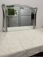 Front angled view of vintage hanging mirror showing its beveled edges and decorative top border reflecting a kitchen and plant.