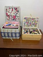 Two vintage sewing baskets displayed side by side on a table. The larger basket on the left has blue and white weaving and an open lid showing sewing notions inside. The smaller basket on the right is natural tone with woven pattern and open lid showing many thread spools inside.