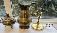 Photo showing three brass items: a lidded container, pitcher, and candlestick holder arranged on a surface by a window.