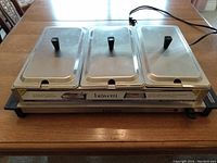 Electric buffet server with three stainless steel chafing dishes with lids on a wooden table, showing power cord and Bravetti brand label