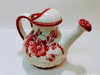 Front side view of the white ceramic watering can with red floral design and trim along handle, spout, and lid.