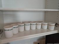Shelf display of all eight Corning Ware mugs, showing two different decorative rim patterns; four with green floral vine, four with beige grapevine.