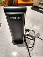 Black OttLite desk lamp standing upright on a counter showing front with logo and power cord coiled beside it.