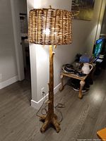Full view of the standing floor lamp showing resin base resembling tree roots and trunk, and woven wicker lampshade illuminated.