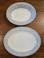 Two oval fine bone china serving pieces with floral patterned blue-gray borders, a meat platter and a vegetable bowl, displayed on wooden surface.