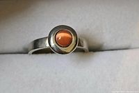 Silver ring with round orange cabochon stone set in bezel, displayed on white cushion.