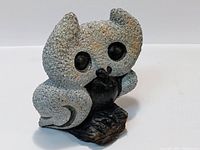 Stone owl sculpture carved from stone, showing textured grey body and black eyes, sitting on dark base resembling a branch.