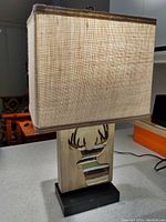 Full lamp showing solid black base, wooden block with carved deer design, and square beige fabric shade
