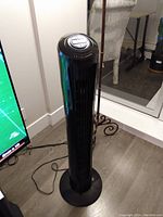 Black tower fan standing on floor near wall, showing full height and overhead control panel.