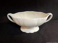 Front angle showing two-handled white ceramic vase with scalloped edges and pedestal base on black background.