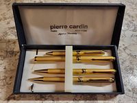 Set of four gold tone ribbed pens and pencil arranged inside open blue Pierre Cardin box showing the pens horizontally side by side with white padded interior and Pierre Cardin branding.