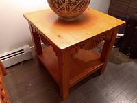 Wood side table positioned against white wall and dark wooden floor with decorative pottery on top. Shows full side and top surfaces with visible wood grain and carved details.
