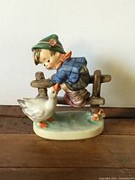 Front view of the Barnyard Hero Hummel figurine showing the boy leaning on a wooden fence with a white goose in front, on an oval green base.