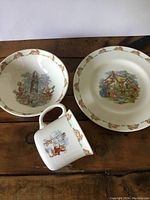 Set of three Bunnykins Royal Doulton items including plate, bowl, and mug on wooden surface showing central bunny scenes and border designs
