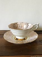 Tea cup and saucer on wooden surface showcasing gold floral detail and white porcelain body.