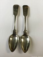Two early 1800s sterling silver spoons with visible inscriptions on the handles, tarnish and patina visible on surface, flat-lay photo on white background.
