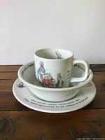 Stacked view of the Peter Rabbit Wedgwood china mug, bowl, and plate all together showing front illustrations and text