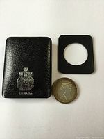 Black leatherette case with silver Canada emblem on cover, smaller black plastic insert for coin display, and silver coin showing profile of Queen Elizabeth II in front of the case.
