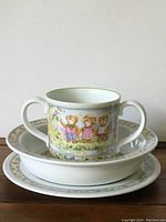 Photo of porcelain loving cup with two handles placed inside matching plate and bowl, showing teddy bear designs and floral border.