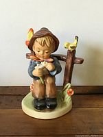 Front view of the Hummel figurine showing the boy seated plucking daisy petals with a yellow bird on the fence.