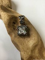 Close-up photo of the sterling silver pendant with square-cut clear crystal, showing detailed setting and bail placed on textured wooden surface.