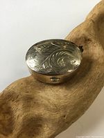 Side angled photo showing oval sterling silver pill box with detailed hand etched floral pattern on lid, placed on a piece of wood.
