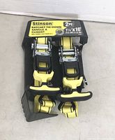 Photo showing two Stinson brand ratchet tie-down straps still wrapped on the original black plastic holder with yellow label detailing size and brand.