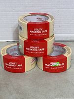Four rolls of beige utility masking tape stacked with red labels showing size and product name.