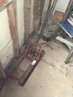Red hydraulic lift with two wooden support platforms and caster wheels, stored in a concrete and wood wall basement corner, visible dust and wear.